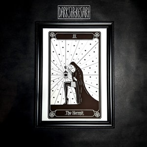 May include: A black and white illustration of The Hermit tarot card. The card features a hooded figure holding a lantern and standing in a field of stars. The card is framed in a black frame with a gold border.