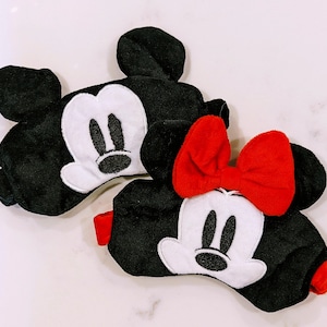 May include: Two black sleep masks with white Mickey Mouse and Minnie Mouse faces. The Minnie Mouse mask has a red bow.