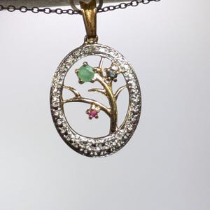 May include: Oval pendant with a tree design. The pendant features a gold-colored tree with green, pink, and blue gemstones. The oval frame is encrusted with small, clear stones. The pendant hangs from a delicate chain.