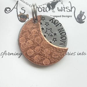 May include: A silver chain necklace with a two-tone pendant. The pendant features a copper crescent moon with a hammered texture and a silver disc with stars and the words "Moon Joy". The necklace is on a white background.