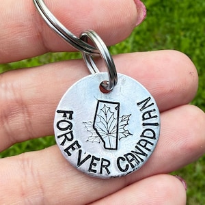 May include: A silver-coloured keychain with a round pendant. The pendant features the words "FOREVER CANADIAN" around the edge and a maple leaf design with the outline of Alberta in the centre. The keychain is held by a person.