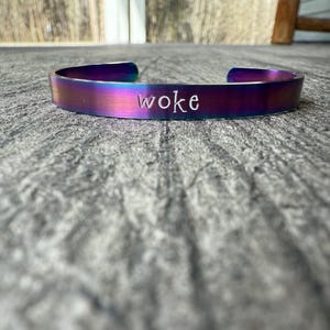 Puede incluir: Una pulsera de metal iridiscente arcoíris con la palabra "woke" grabada en ella.