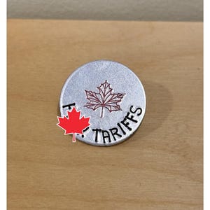 May include: A circular, silver-coloured metal object featuring the word "TARIFFS" in black. A red maple leaf design is in the centre, with a smaller maple leaf graphic in the lower left corner.