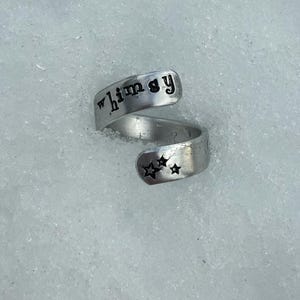 May include: A silver-colored wrap ring with the word "whimsy" stamped on the top band. The bottom band features three stamped stars. The ring is set against a white, textured background.