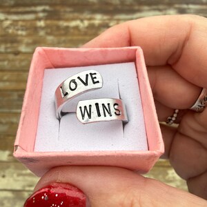 May include: A silver-colored ring with the words "LOVE WINS" engraved on it, presented in a small pink box. The ring has an open design, with the words split across the two ends. The ring is displayed against a light pink background.