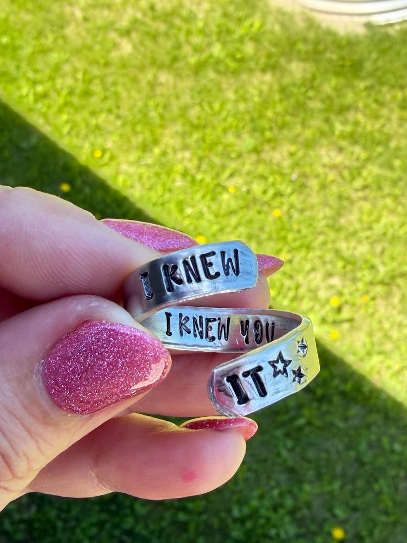 Puede incluir: Un anillo de metal plateado con dise&ntilde;o envolvente que presenta el texto negro grabado I KNEW I KNEW YOU IT junto a dos peque&ntilde;os s&iacute;mbolos de estrellas. El anillo tiene un dise&ntilde;o abierto y ajustable con un acabado liso y pulido.