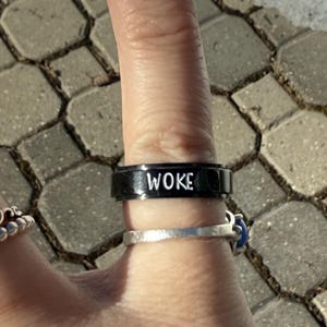 Könnte beinhalten: Ein schwarzer Ring mit der Aufschrift "WOKE" in weißen Buchstaben.