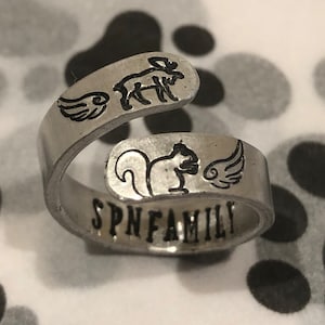 Könnte beinhalten: Ein silberner Ring mit einem Eichhörnchen, einem Elch und zwei Flügeln, die darauf graviert sind. Der Ring trägt auch die Worte "SPN FAMILY", die darauf graviert sind.