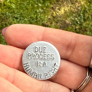 Pode incluir: Um token de metal prateado com o texto "Due Process Is A Human Right" gravado nele.
