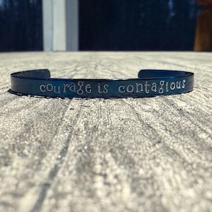 May include: A dark blue metal cuff bracelet with the words "courage is contagious" engraved in a cursive font. The bracelet is resting on a textured, light gray surface, with a blurred background.