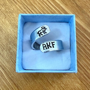 Może przedstawiać: Srebrny pierścionek z napisem "AKF" na jednym końcu i małą sylwetką zwierzęcia na drugim. Pierścionek jest prezentowany w małym, jasnoniebieskim pudełku z białym wnętrzem.