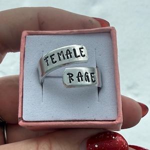 Puede incluir: Un anillo plateado con las palabras "FEMALE" y "RAGE" estampadas. El anillo está en una caja rosa con acolchado blanco. El anillo es de estilo envolvente.