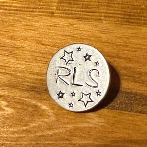 May include: A small, silver metal disc featuring the initials "RLS" in a handwritten style, surrounded by star shapes. The disc rests on a wooden surface. The disc's diameter is approximately 2.5 cm.