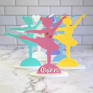 May include: A set of five ballerina earring display stands in various colors, including pink, blue, yellow, and white. The stands are made of acrylic and feature a ballerina silhouette with a hole for earrings. The stand in the center has the name "Quinn" written on it.