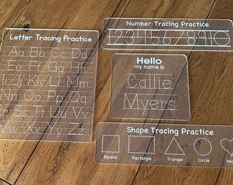 Peut inclure: Quatre planches de traçage en acrylique transparent pour enfants. Les planches présentent des lettres majuscules, des chiffres, des formes et une section de traçage de nom avec le texte "Hello my name is".