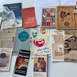 Könnte beinhalten: Eine Sammlung von Vintage-Kochbüchern mit bunten Umschlägen. Die Kochbücher tragen Titel wie "Menu Magic", "Holiday Inn Cookbook", "What Makes Jell?" und "Choice Recipes North Atlantic Sea Foods".