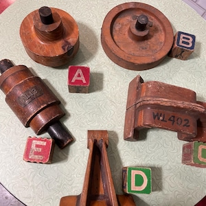 May include: A collection of vintage wooden objects and alphabet blocks. The wooden items include a wheel-shaped piece, a cylindrical piece, and a frame-like structure. The alphabet blocks are painted with the letters A, B, C, D, and E. The items are arranged on a light green table.