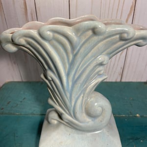 Könnte beinhalten: Eine hellblaue Keramikvase mit einem einzigartigen, stilisierten Design. Die Vase hat eine ausgestellte, fächerartige Form mit einem wirbelnden, von Muscheln inspirierten Motiv. Der Sockel hat eine rechteckige Form. Der obere Rand hat einen rosafarbenen Farbton.