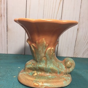 May include: A brown and green ceramic vase with a unique, stylized design. The vase has a wide, flared opening and a base that resembles a curled leaf.