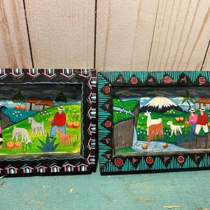 May include: Two rectangular wooden shadow boxes featuring hand-painted Andean landscapes. The left frame is black with pink accents, showing a person herding sheep. The right frame is teal with red accents, depicting llamas and people near a waterfall.