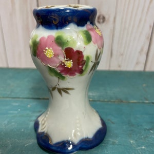 May include: A small white ceramic vase with a blue and gold rim. The vase is decorated with pink and red flowers and green leaves.