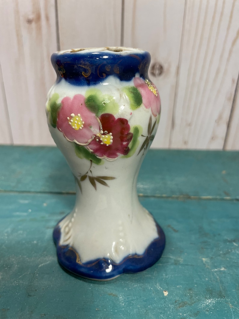 May include: A small white ceramic vase with a blue and gold rim. The vase is decorated with pink and red flowers and green leaves.