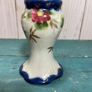 May include: A white ceramic candle holder with a blue and gold floral design. The holder has a wide base and a narrow neck.