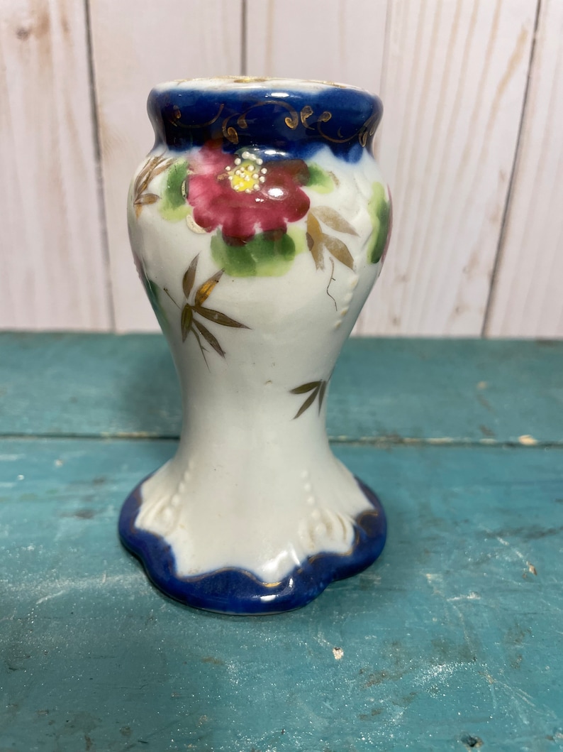 May include: A white ceramic candle holder with a blue and gold floral design. The holder has a wide base and a narrow neck.