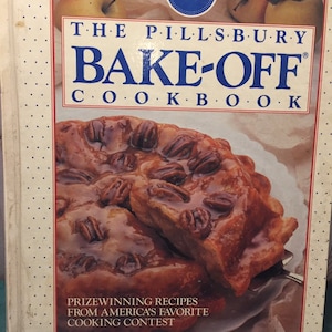 Könnte beinhalten: Das Pillsbury Bake-Off Kochbuch mit einem Kuchen auf dem Cover. Das Kochbuch hat einen weißen Einband mit rotem und blauem Text. Das Cover zeigt eine Nahaufnahme eines Pekannusskuchens. Der Text lautet "Preisgekrönte Rezepte aus Amerikas beliebtestem Kochwettbewerb."
