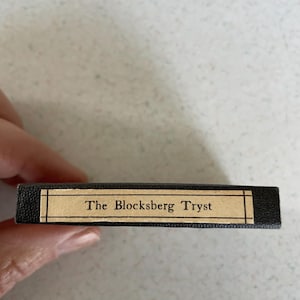 May include: A small, rectangular black box with the text "The Blocksberg Tryst" on a tan label. The box is held in a person's hand, with a light-coloured surface in the background.