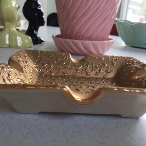 May include: A white ceramic ashtray with a gold rim and a textured gold surface. The ashtray has a square shape with rounded corners and a slight indentation in the center.