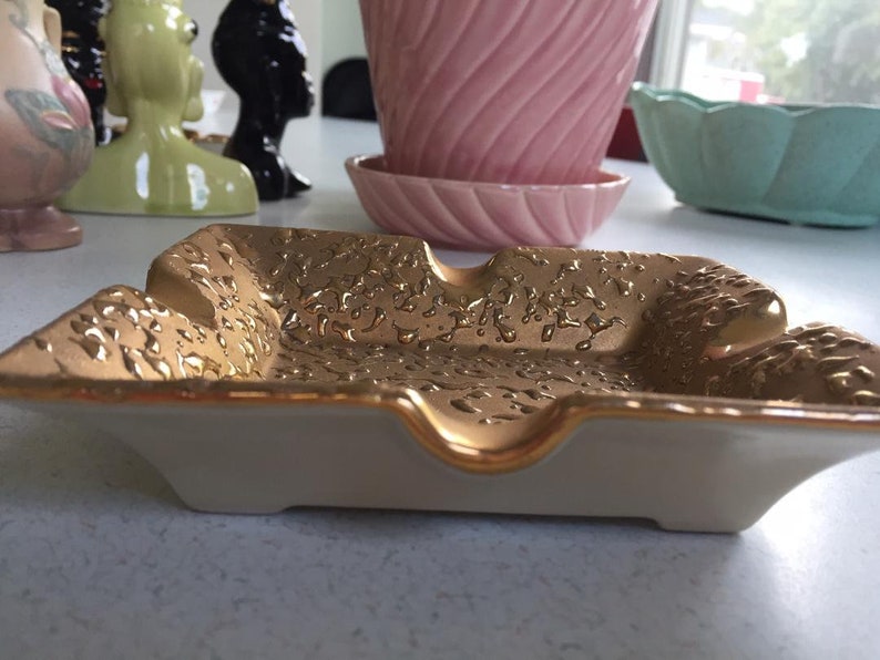 May include: A white ceramic ashtray with a gold rim and a textured gold surface. The ashtray has a square shape with rounded corners and a slight indentation in the center.