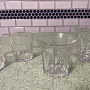 May include: Four clear glass drinking glasses with different shapes and patterns. The glasses are on a green and white patterned surface.