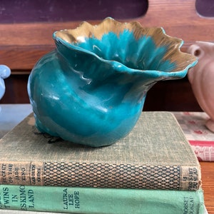 Puede incluir: Un jarrón de cerámica de color verde azulado con un borde dorado. El jarrón tiene forma de concha y un borde ondulado. El jarrón está colocado sobre una pila de tres libros.