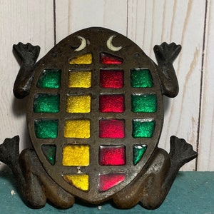 May include: A cast iron frog figurine with a stained glass design in shades of green, red, and yellow. The frog has a whimsical expression with white eyes and a raised tail.