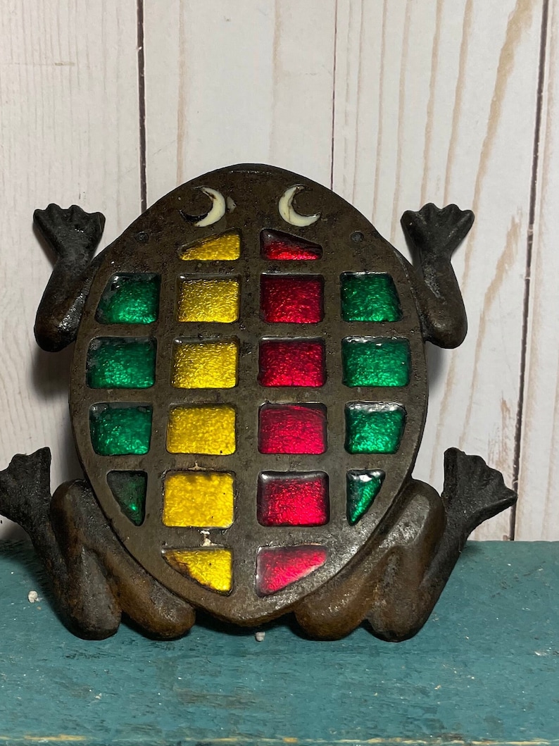 May include: A cast iron frog figurine with a stained glass design in shades of green, red, and yellow. The frog has a whimsical expression with white eyes and a raised tail.
