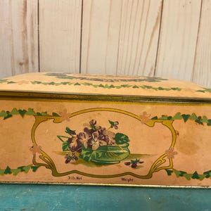 May include: Vintage rectangular tin box with a peach background. The box features a floral design with purple violets in a green bowl, framed by a gold and green border. The top of the box has a floral design. The box weighs 3 lbs.