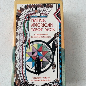 Puede incluir: Una caja de tarot vintage con el título "NATIVE AMERICAN TAROT DECK". La caja presenta ilustraciones de inspiración nativa americana, incluyendo un tocado y un atrapasueños. También es visible el texto "Complete with Booklet of Instructions".
