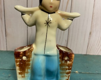 Puede incluir: Una figurita de cerámica de una persona que lleva dos cestas sobre sus hombros. La persona lleva una camisa blanca con un diseño de estrella, pantalones azules y tiene el pelo negro. Las cestas son marrones con detalles blancos.