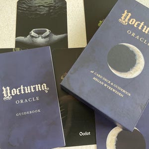 Könnte beinhalten: Ein dunkelblaues Orakelkarten-Deck und ein Handbuch-Set. Die Box und das Handbuch tragen den Titel "Nocturna Oracle" in kunstvollen Buchstaben. Die Box zeigt eine Halbmond-Grafik. Das Deck enthält 48 Karten und stammt von Megan Wyreweden.