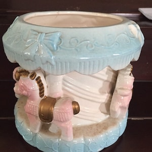 Könnte beinhalten: Ein Keramik-Pflanztopf in Form eines Karussells mit zwei rosafarbenen Pferden. Der Topf ist mit blauen und weißen Details verziert und hat ein weißes Inneres.