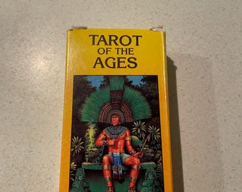 May include: A rectangular box of "Tarot of the Ages" cards. The box is yellow with a colorful illustration of a seated figure in a headdress. The figure is holding a staff. The box has the title in large black letters.