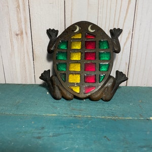 May include: A cast iron frog with a stained glass design in shades of green, red, and yellow. The frog is sitting on a blue surface.