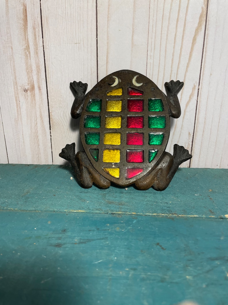May include: A cast iron frog with a stained glass design in shades of green, red, and yellow. The frog is sitting on a blue surface.