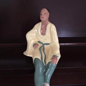 May include: A ceramic figurine of a person wearing a yellow robe with a blue sash and blue pants. The figurine is sitting with its legs crossed and has a neutral expression on its face.