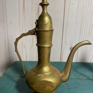 May include: A brass teapot with an ornate design and a hinged lid. The teapot has a spout and a handle. The teapot is on a green surface.