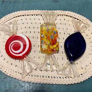 Könnte beinhalten: Drei Glas-Bonbon-Skulpturen auf einer cremefarbenen, ovalen Häkeldecke. Die Bonbons sind rot-weiß-Wirbel, gelb mit bunten Flecken und tiefblau. Jedes ist in durchsichtigem Zellophan verpackt.