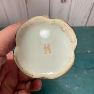 May include: The underside of a small, white ceramic vase with a blue and orange floral design. The vase has a scalloped rim and a flat base. The words "Made in Japan" are printed in orange on the bottom.