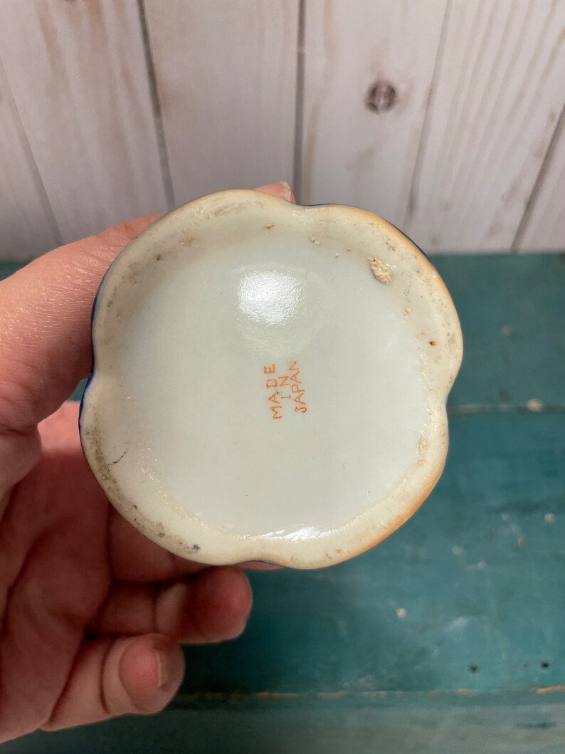 May include: The underside of a small, white ceramic vase with a blue and orange floral design. The vase has a scalloped rim and a flat base. The words "Made in Japan" are printed in orange on the bottom.