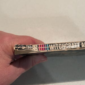 K&ouml;nnte beinhalten: Buchr&uuml;cken mit dem Titel "WITCHCRAFT" in wei&szlig;en Buchstaben. Der R&uuml;cken hat eine Reihe vertikaler Streifen in Rot, Rosa, Blau und Lila. Die Kanten des Buches weisen Gebrauchsspuren auf.